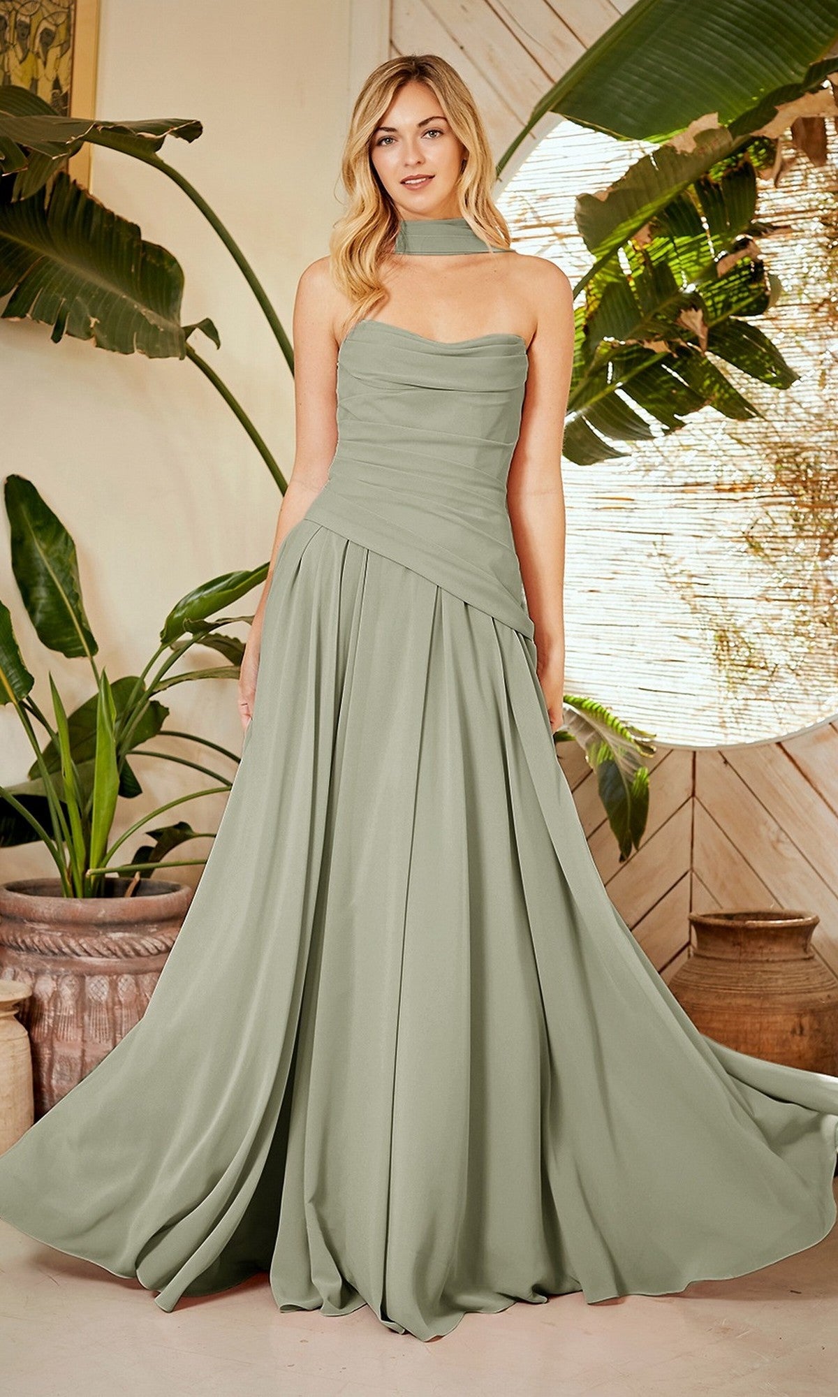 A woman stands indoors surrounded by large potted plants, wearing the Amelia Couture 6133 long prom dress in sage with a strapless neckline, corset back, and flowing chiffon skirt.